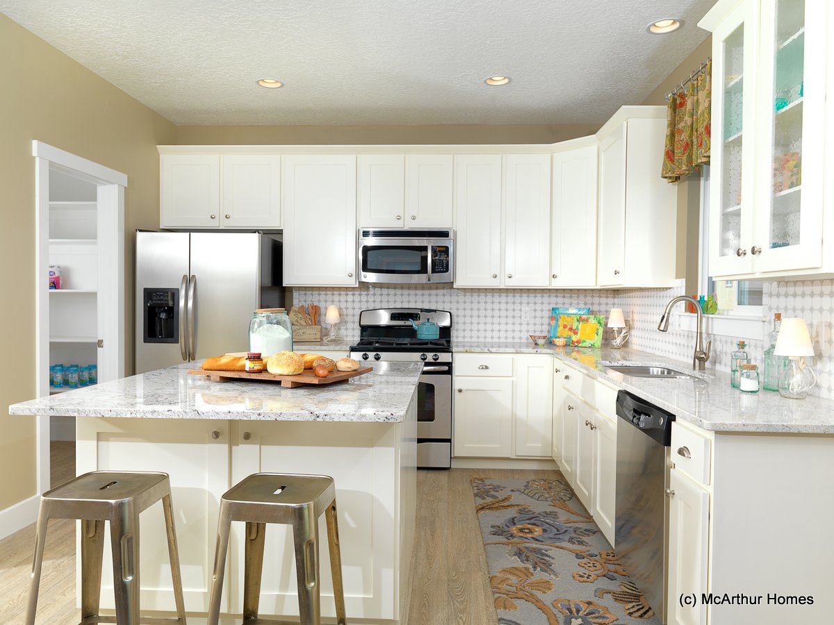 Modern kitchen with white cabinets, stainless steel appliances, marble island with stools, and baked goods on the counter. Pantry and refrigerator are visible in the background—a bright, clean space typical of homes in Utah.