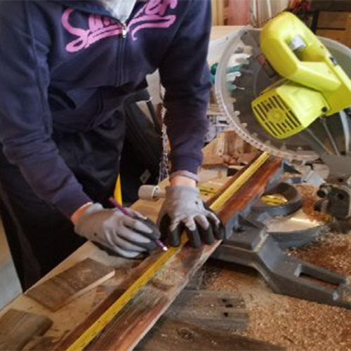 Person wearing gloves measuring a reclaimed wood plank with a tape measure and marking it with a pencil, next to a yellow miter saw on a workbench covered in sawdust.