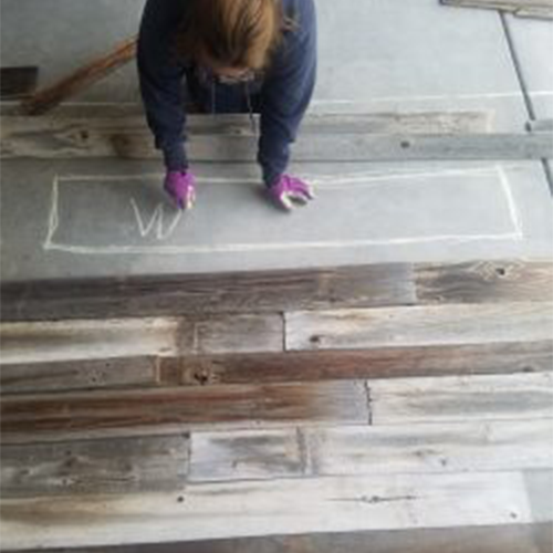 Person wearing purple gloves marking a rectangular outline with chalk on a reclaimed wood surface.