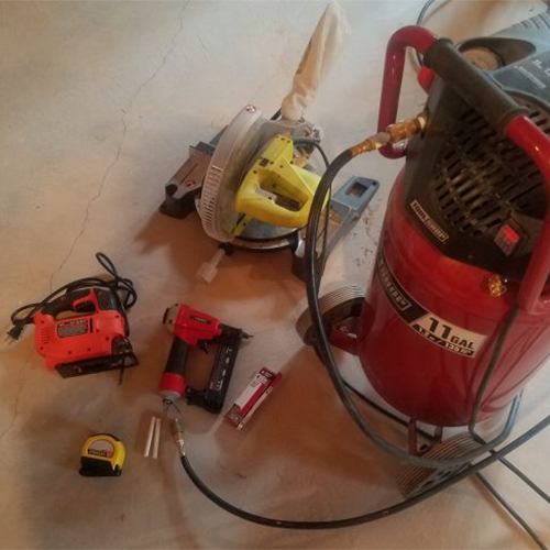 Collection of construction tools on a concrete floor, including a red air compressor, a yellow circular saw, a red nail gun, a drill, a tape measure, and other small tools.