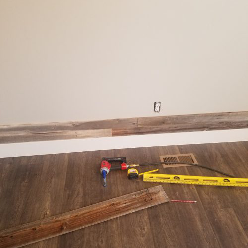 Partially installed reclaimed wood wall with tools on the floor, including a drill, tape measure, level, and a piece of wood plank, in a room with dark wood flooring.