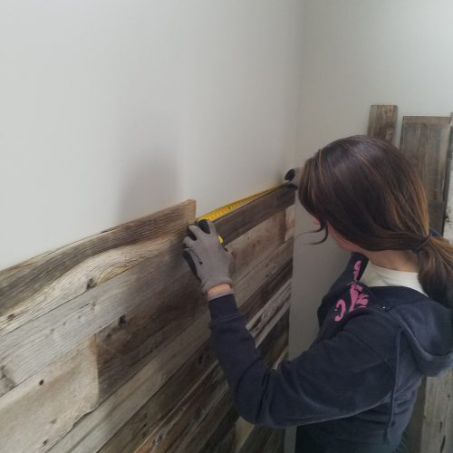 Woman wearing gloves measuring reclaimed wood planks on a wall with a yellow measuring tape.