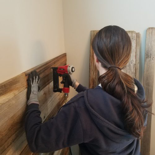 Woman wearing gloves using a nail gun to attach reclaimed wood planks to a wall.