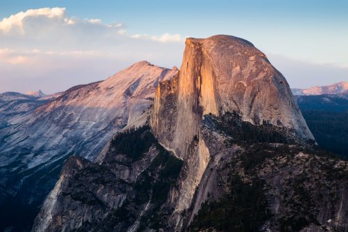 Sunlight illuminates the sheer granite face of Half Dome in Yosemite National Park, reminiscent of landscapes near homes in Utah, with rugged, forested mountains stretching into the distance under a partly cloudy sky.