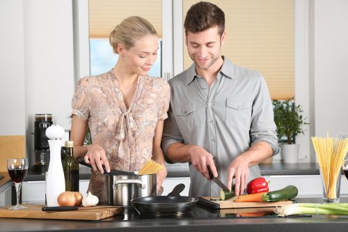 Smiling couple cooking together in a modern kitchen, with the woman placing spaghetti into a pot and the man chopping vegetables on a cutting board.