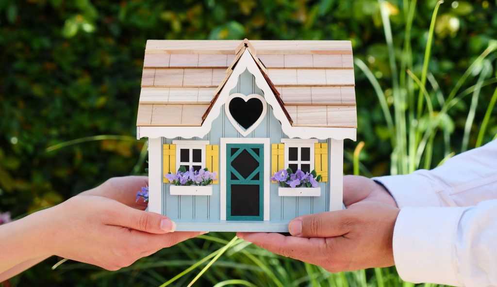 2 People Holding A Small Wooden House With A Heart in the Middle