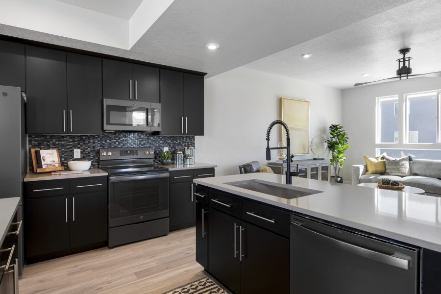 Contemporary kitchen with black cabinets, stainless steel appliances, a mosaic tile backsplash, and a large island with a modern black faucet, opening into a bright living area with a sectional sofa and large windows.