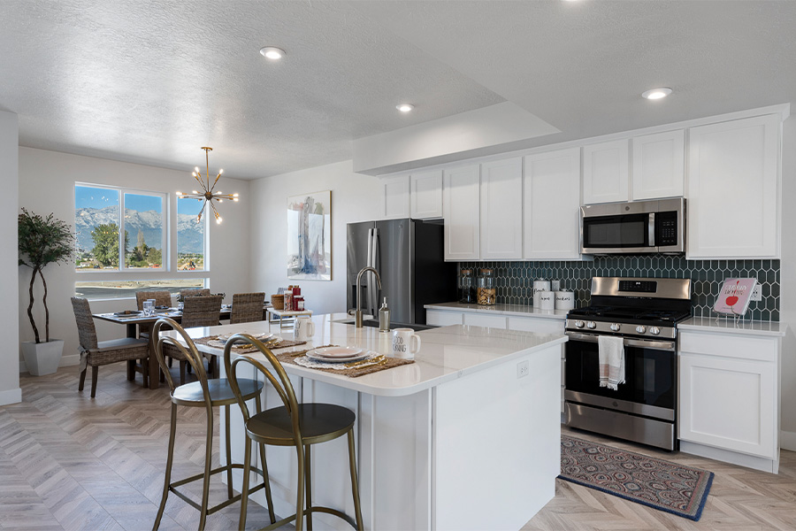 Modern open-concept kitchen and dining area with white cabinetry, stainless steel appliances, a black hexagonal tile backsplash, and a large island with seating for two. Dining table with woven chairs is set near a window with a mountain view.