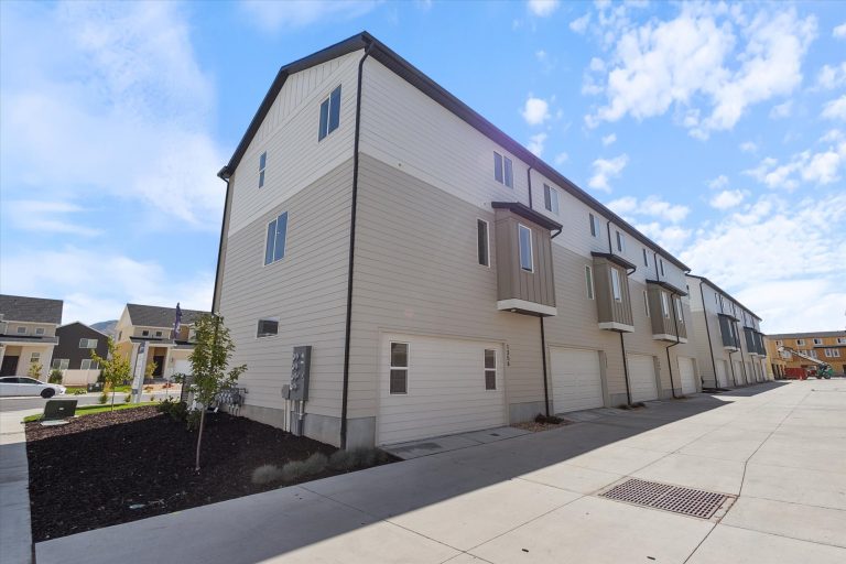 A row of modern, three-story townhouses with beige siding and white trim, each with a garage. The pavement is clean, and the sky is mostly clear with some scattered clouds.