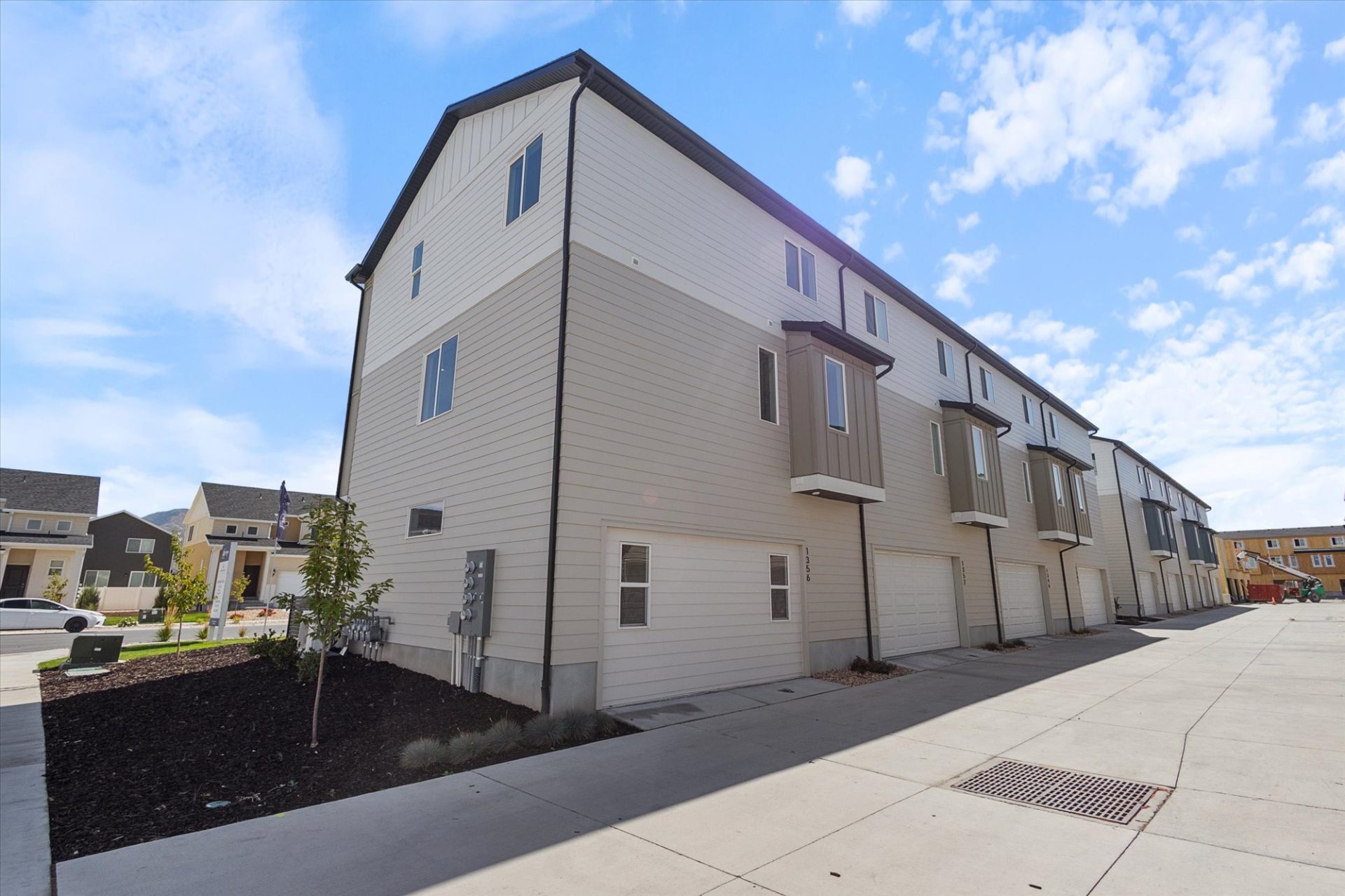 A row of modern, three-story townhouses with beige siding and white trim, each with a garage. The pavement is clean, and the sky is mostly clear with some scattered clouds.