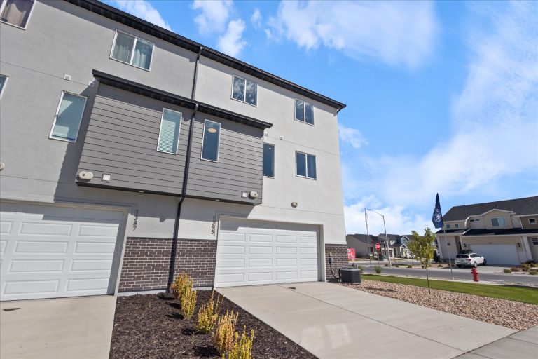 A modern three-story townhouse with a gray and white exterior, two garage doors, and small shrubs planted in front. The sky is bright blue with scattered clouds, and neighboring houses are visible.