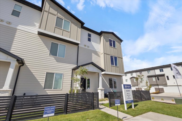 Modern three-story townhouse with beige and white siding, large windows, and a small fenced front yard. A sign near the entrance displays model home information. Blue sky and another similar building are visible.