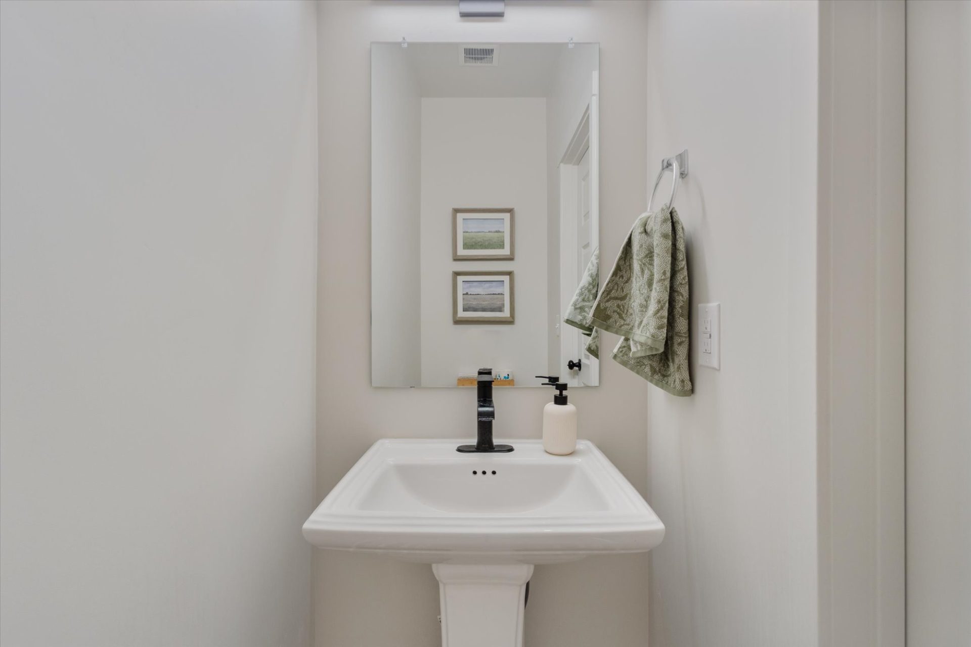 A small, minimalist bathroom with a white pedestal sink, a rectangular mirror above it, a soap dispenser, a towel hanging on the right wall, and two landscape photos reflected in the mirror.