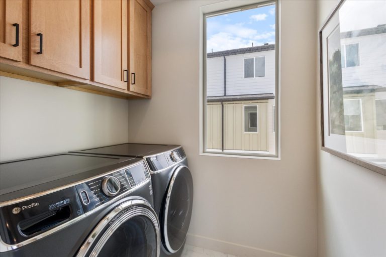 A modern laundry room with a front-loading washer and dryer, wooden cabinets above, a window letting in natural light, and a framed picture on the wall.