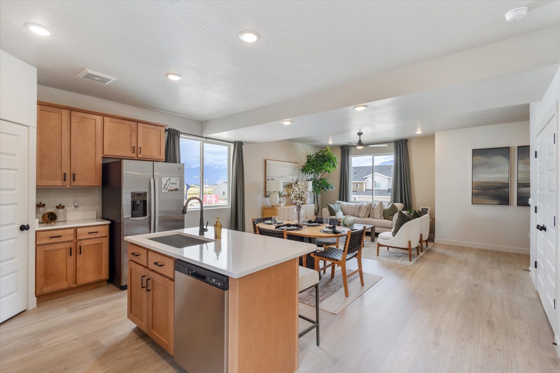 Modern open-concept kitchen and living room with light wood floors, wood cabinets, stainless steel appliances, island with sink, dining table, large windows, and neutral décor with plants and artwork.