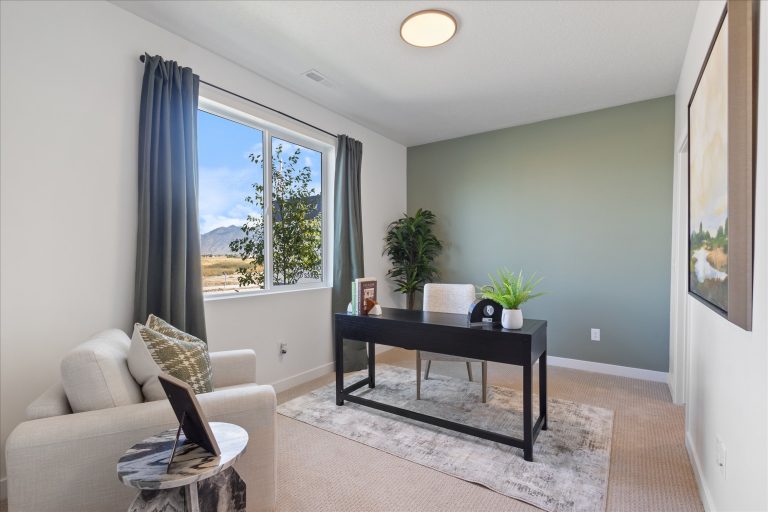 A modern home office with a desk, chair, and laptop, a potted plant, and an armchair near a window with mountain views. The room has light walls, a green accent wall, and a large artwork.
