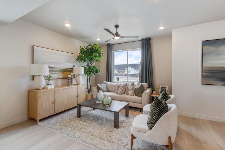 Modern living room with a neutral palette, featuring a beige sofa, accent chair, wooden coffee table, sideboard with lamps and decor, large potted plant, window with gray curtains, and framed artwork on the walls.