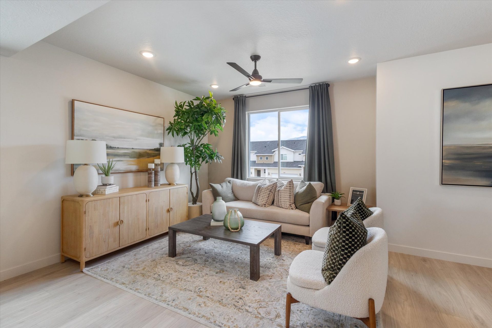 Modern living room with a neutral palette, featuring a beige sofa, accent chair, wooden coffee table, sideboard with lamps and decor, large potted plant, window with gray curtains, and framed artwork on the walls.
