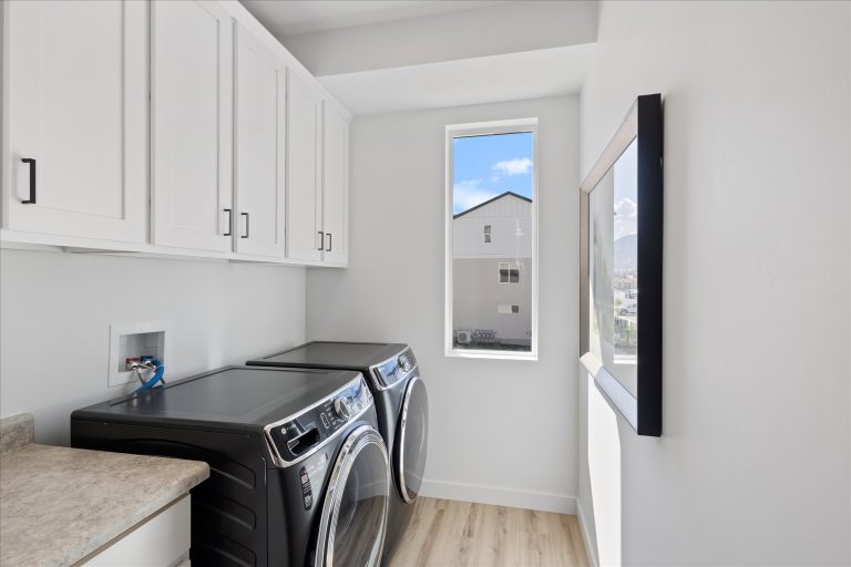 A bright laundry room with white cabinets, a countertop, and a black washer and dryer set, featuring a window with an outdoor view and framed art on the wall.