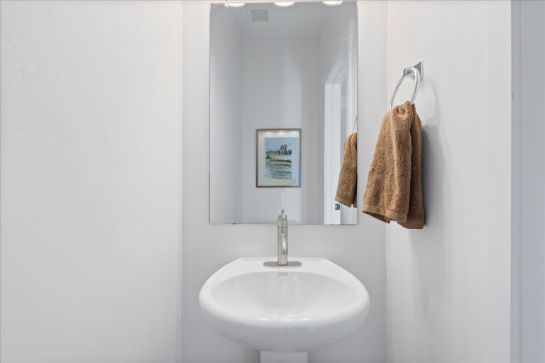 A modern white bathroom features a pedestal sink, a mounted mirror, two brown hand towels on a ring holder, and a framed artwork reflected in the mirror. The space is minimalistic and well-lit.