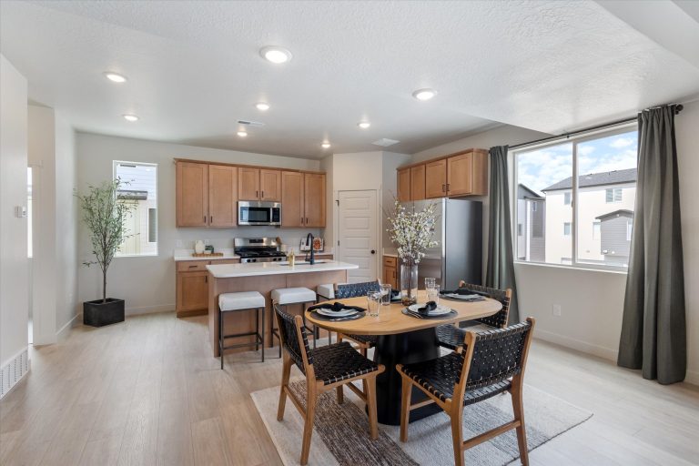 Modern kitchen and dining area with wood cabinets, stainless steel appliances, a round dining table set for four, woven chairs, light wood flooring, large windows, and natural light.