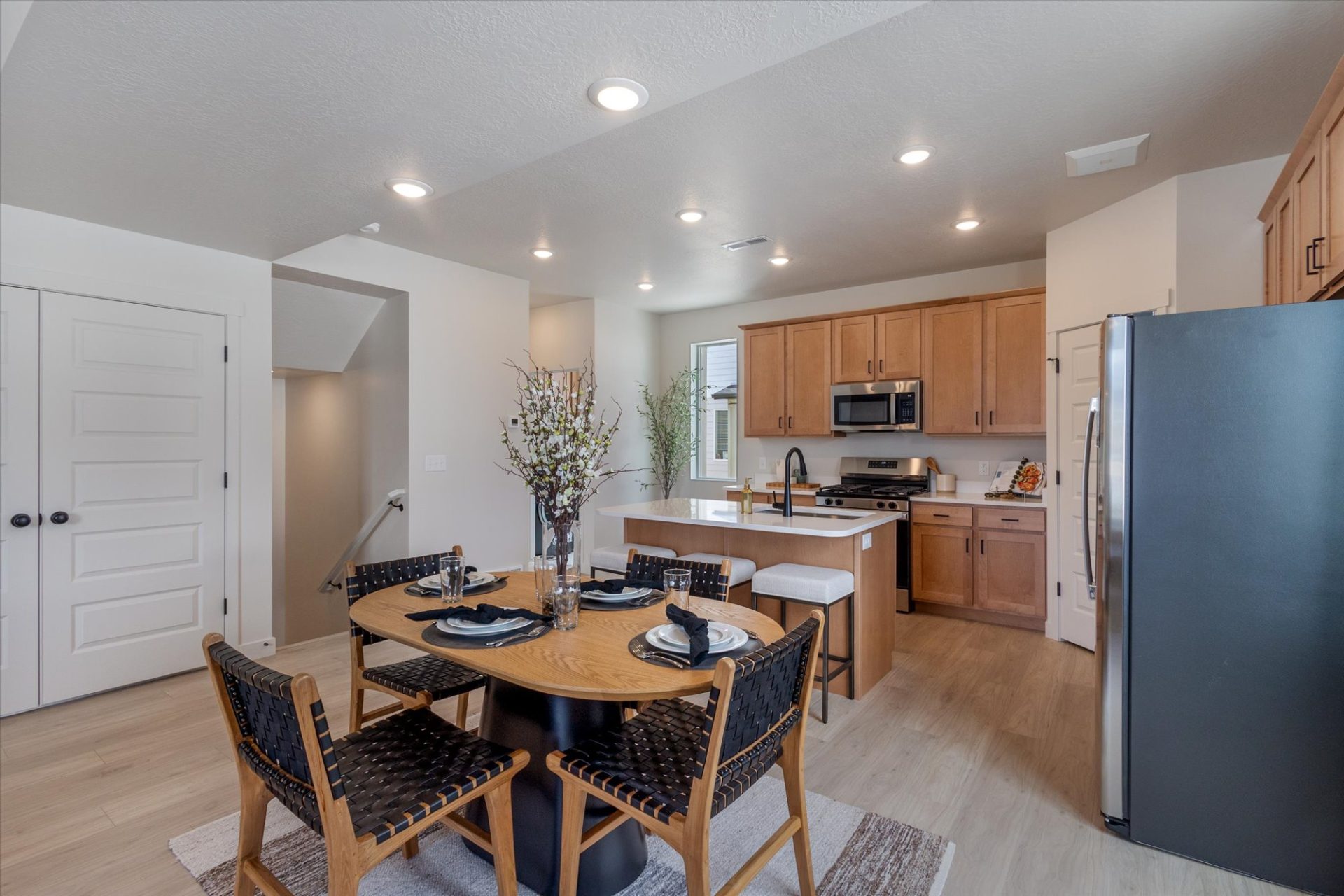 A modern kitchen and dining area with light wood floors, wooden cabinets, stainless steel appliances, a round dining table set for four, and a centerpiece of tall branches in a vase.