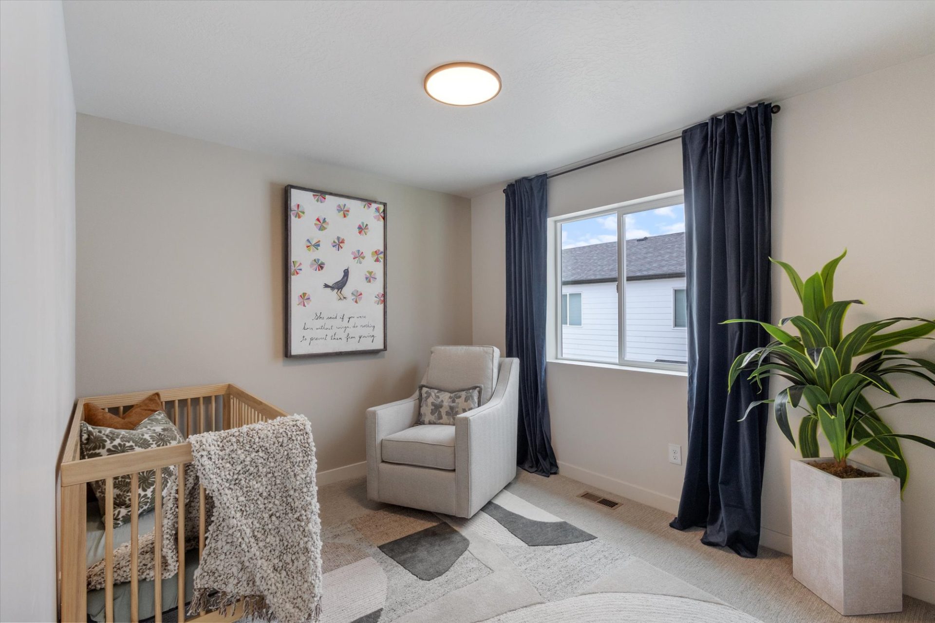 A cozy nursery with a wooden crib, a cushioned gray armchair, a large leafy plant, dark curtains, a window letting in natural light, and a framed, colorful butterfly print on the wall.