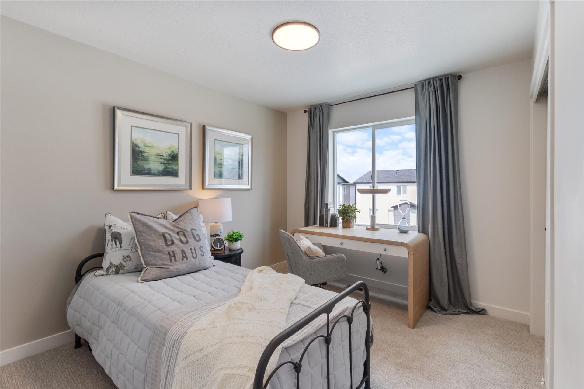 A cozy bedroom with a single bed, gray bedding, and decorative pillows, including one that says DOG HAUS. A desk with a chair sits by a window with gray curtains, letting in natural light. Art hangs on the beige wall.