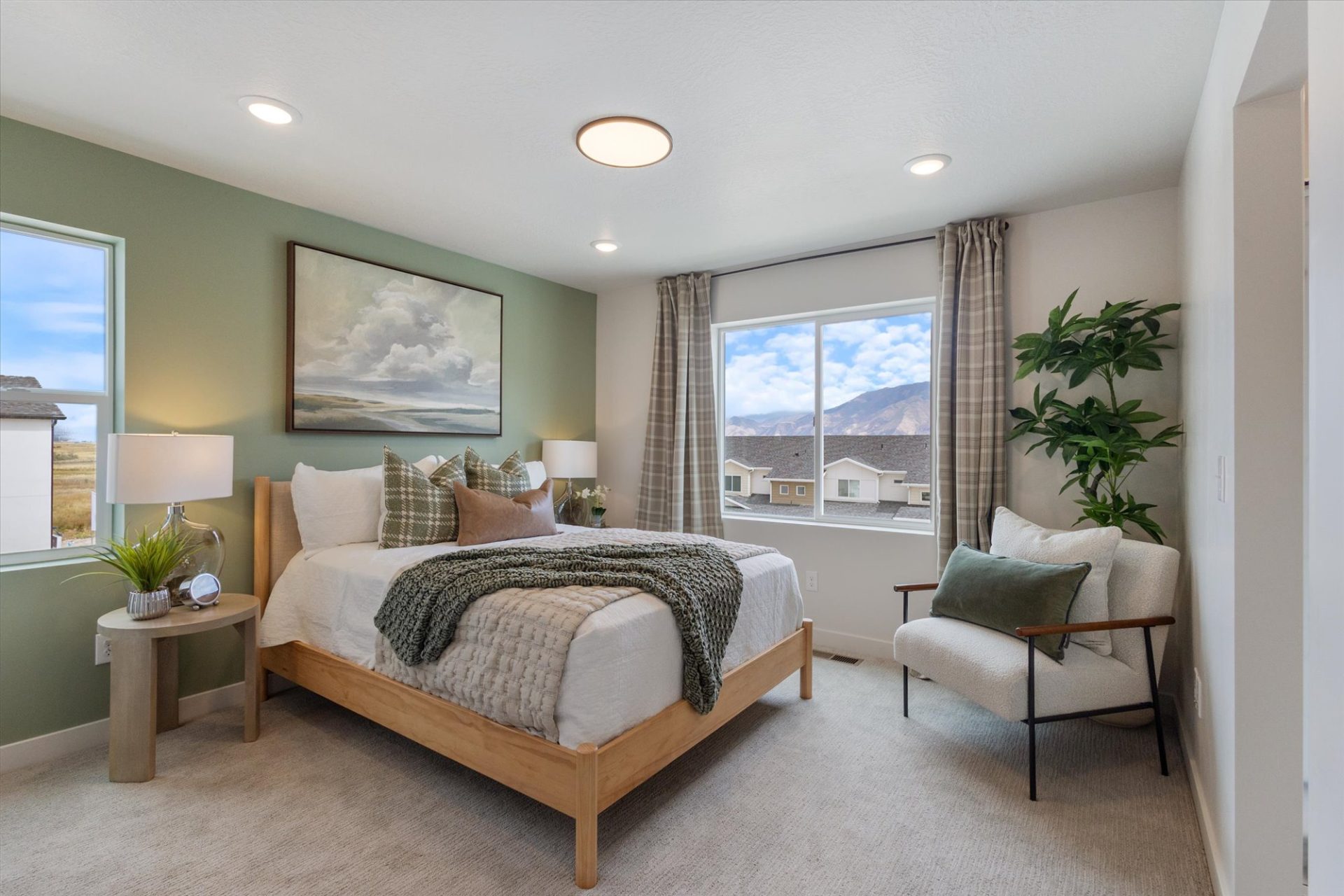 A bright, modern bedroom with a wooden bed, white bedding, green accent wall, large landscape painting, two windows with mountain views, a cozy chair, and a potted plant. Neutral decor and soft lighting complete the space.