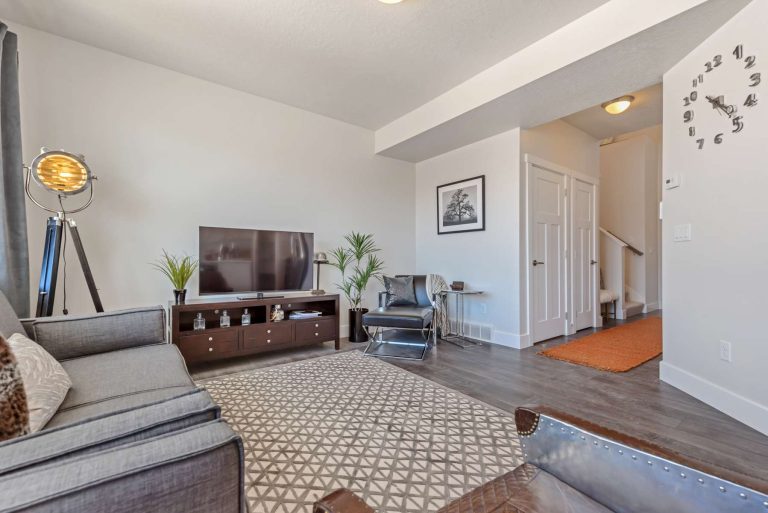 Modern living room with gray sofa, TV on a wooden stand, potted plants, tripod lamp, and patterned rug—features often found in stylish homes in Utah. White walls and doors, hardwood floors, and a staircase complete the inviting space.