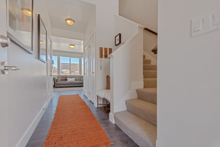 A bright hallway with dark wood floors, white walls, and an orange runner rug welcomes you into one of the stunning homes in Utah. The space leads to a living area with large windows, a gray sofa, and a stairway with beige carpet.