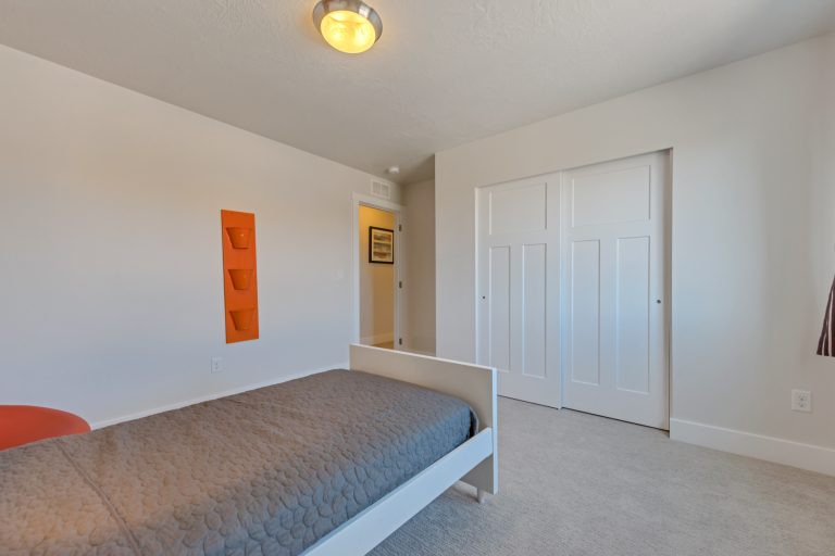 Minimalist bedroom with beige walls, a single bed with a gray quilt, orange wall storage bins, double white closet doors, and a small ceiling light fixture—perfect for those seeking bright, simple spaces often found in homes in Utah.
