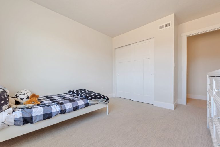 Minimalist bedroom in homes in Utah with beige carpet, light walls, a single bed with black-and-white bedding, stuffed animals, a dresser, and a double closet with white sliding doors. The room appears bright and tidy.
