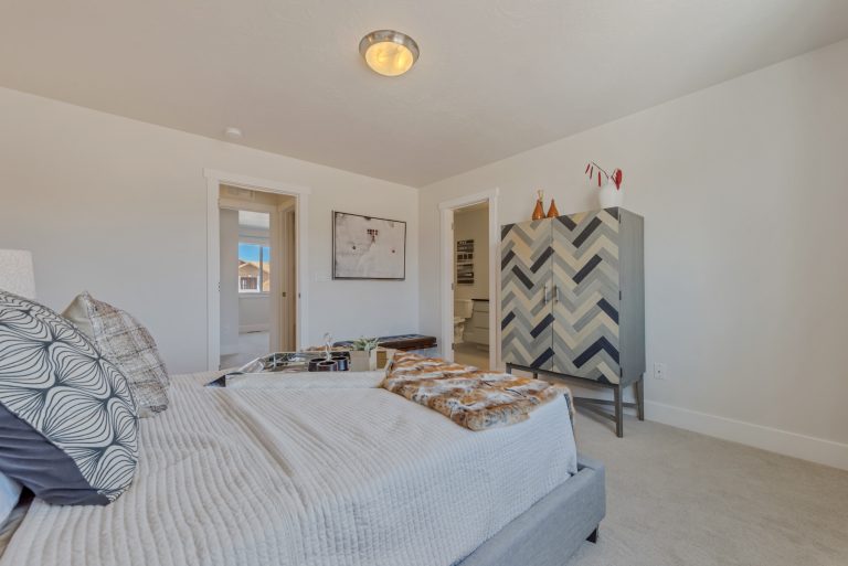 A modern bedroom in homes in Utah features a neatly made bed, decorative pillows, a chevron-patterned wardrobe, light walls, carpet flooring, and doorways to a bathroom and hallway. Natural light streams in from the window.