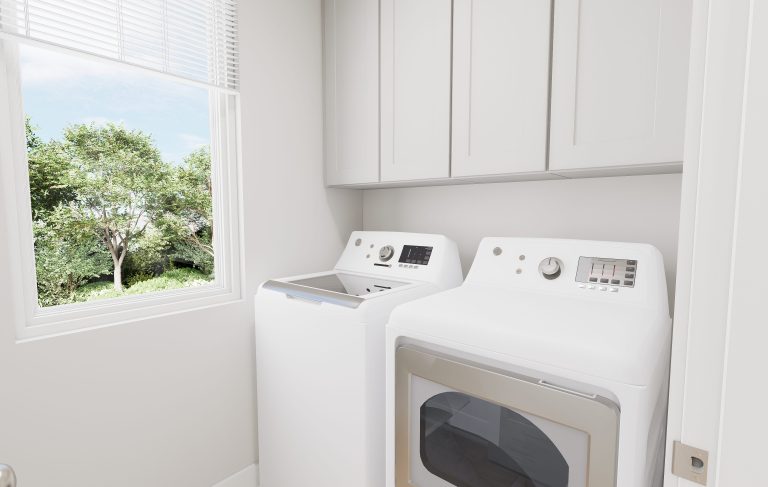 A bright laundry room in homes in Utah, with a window overlooking green trees, features a white top-loading washing machine and a white front-loading dryer beneath sleek white cabinets.