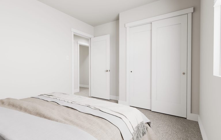 A minimalist bedroom found in many homes in Utah, featuring a neatly made bed, beige and white bedding, light carpet, white walls, a door, double sliding closet doors, and soft natural light streaming through a window on the right.