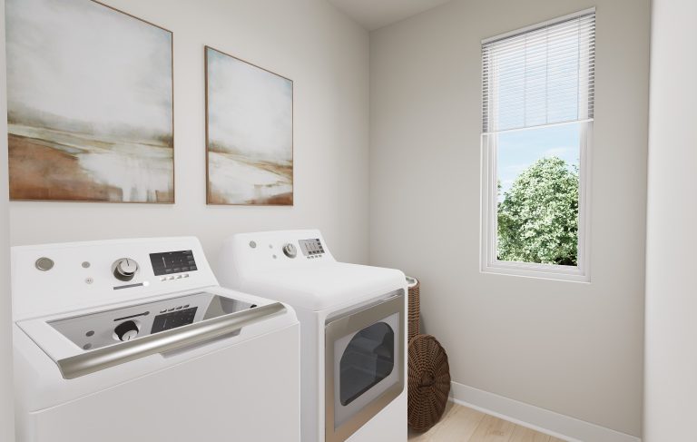 A modern laundry room found in many homes in Utah, featuring a white washer and dryer, two abstract paintings on the wall, a sunlit window, and a wicker basket in the corner.