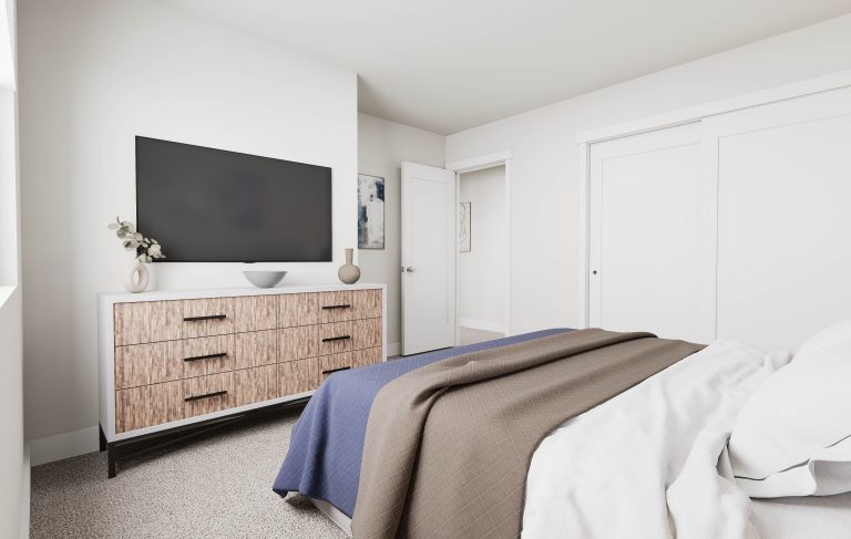 A modern bedroom in homes in Utah, featuring a neatly made bed with blue and brown blankets, a wooden dresser topped with a large TV, light carpeting, and white walls adorned with minimal decor.