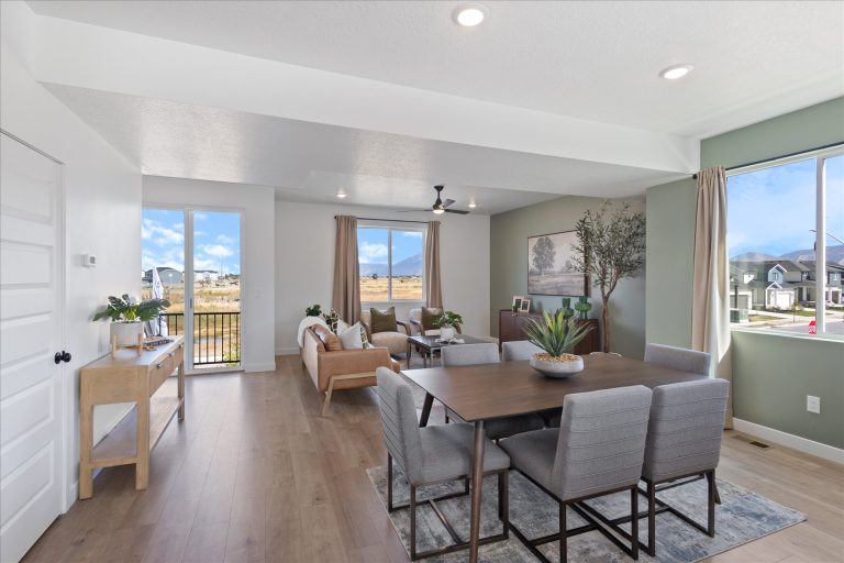 Modern open-concept living and dining area with large windows, neutral decor, gray chairs around a dining table, light wood flooring, and natural light filling the space. Houses and mountains visible outside.