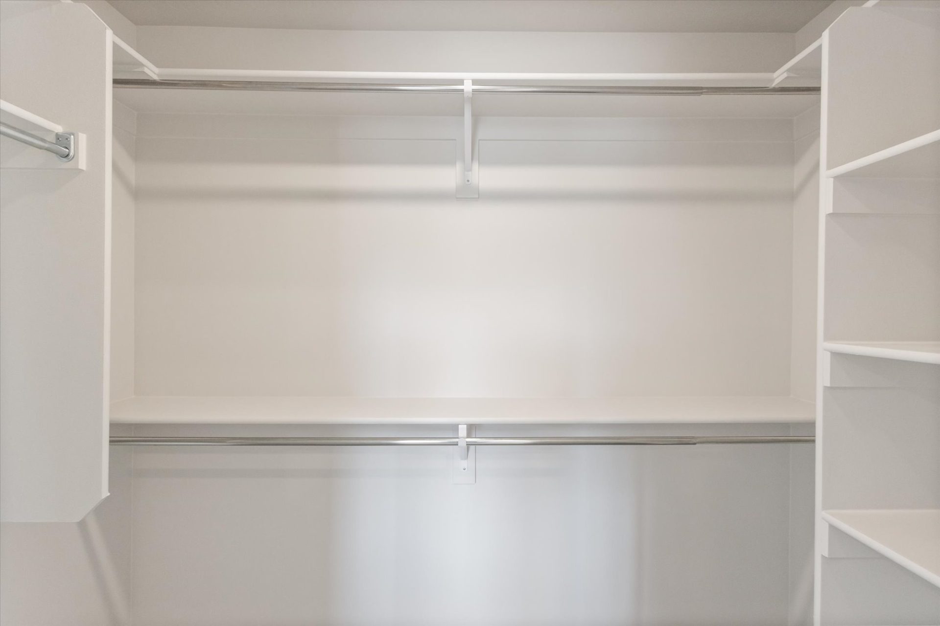 Empty walk-in closet with white walls, two metal hanging rods, a high shelf above the rods, and open shelving units on the right side. The closet is clean and well-lit.