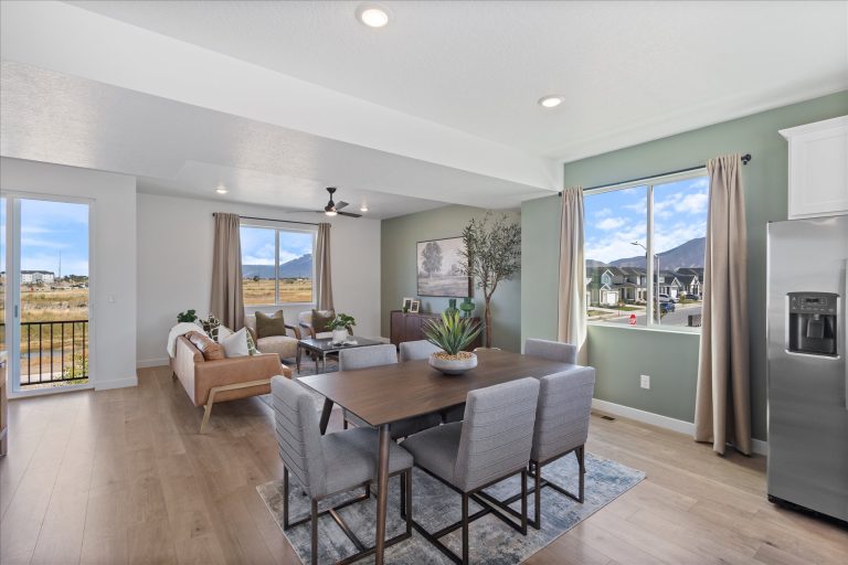 Modern open-concept living and dining area with large windows, mountain views, a wooden dining table with six gray chairs, a light sofa, and contemporary decor in neutral tones.