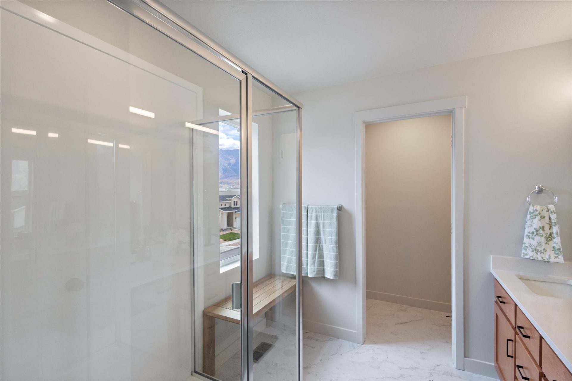 Modern bathroom with a glass-enclosed shower, a wooden bench inside, a towel hanging on a rack, light-colored walls and floor, and a vanity with a floral hand towel. A window reveals a view outside.