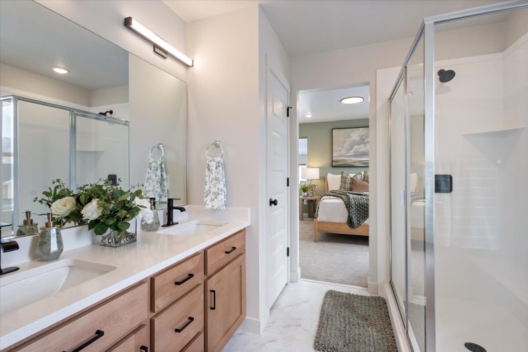 Modern bathroom with double sinks, wood cabinets, large mirror, glass shower, towel racks with patterned towels, and a vase of flowers. A doorway opens to a bright, neatly made bedroom with a wooden bed.