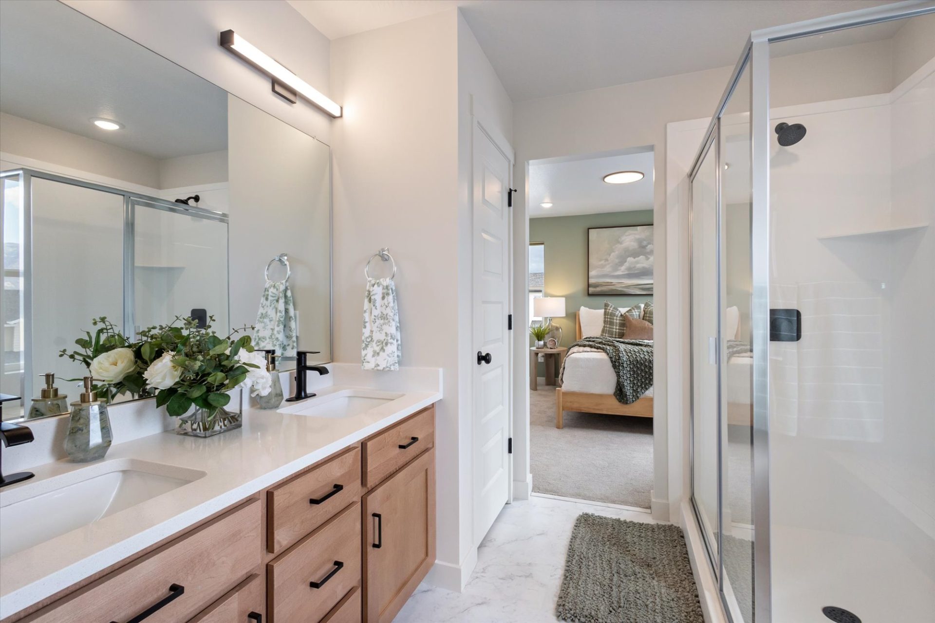 Modern bathroom with double sinks, wood cabinets, large mirror, glass shower, towel racks with patterned towels, and a vase of flowers. A doorway opens to a bright, neatly made bedroom with a wooden bed.