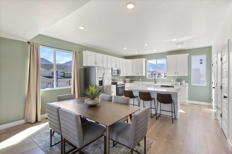 Modern kitchen and dining area with light green walls, white cabinets, stainless steel appliances, a wooden dining table with six upholstered chairs, a kitchen island with three barstools, large windows, and wood flooring.