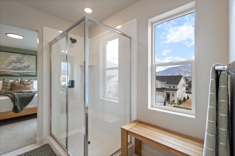 Modern bathroom with a glass shower enclosure, a wooden bench, and a towel hanging on a rack; large windows let in natural light and reveal a view of houses and mountains outside. Bedroom visible through open doorway.
