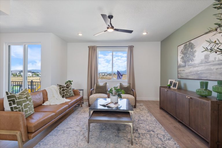 A modern living room with a tan leather sofa, two armchairs, a coffee table, and a green accent wall. Large windows let in natural light, and decor includes plants, artwork, and green vases.