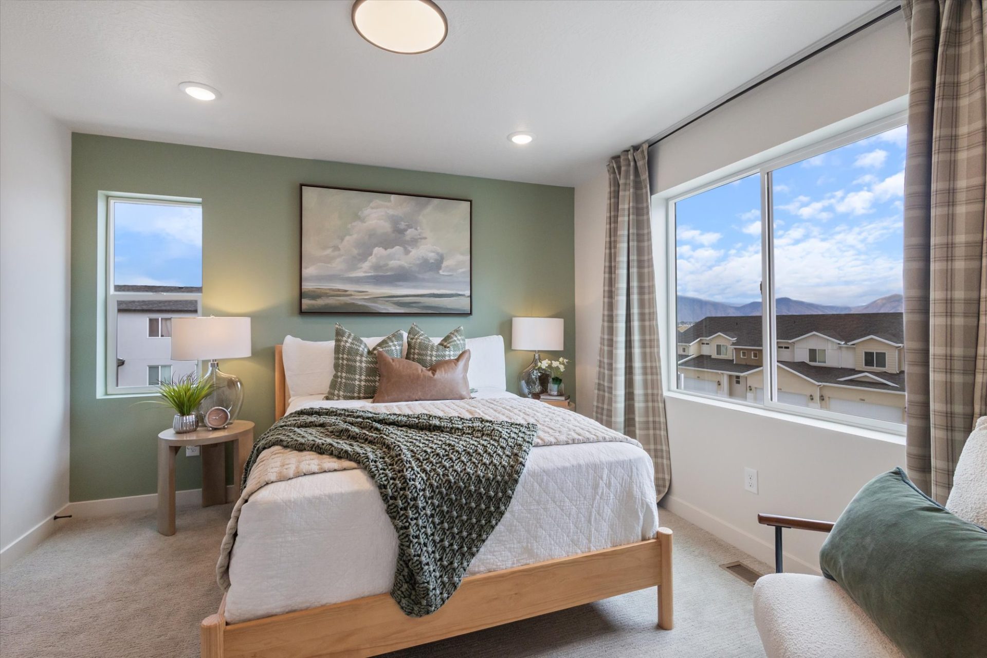 A cozy bedroom with a wooden bed, neutral bedding, and green pillows. Large windows let in natural light, offering views of neighboring houses and mountains. A landscape painting hangs above the bed, and two lamps sit on bedside tables.