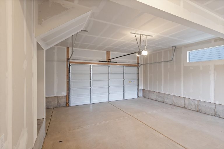 A clean, empty two-car garage with unfinished drywall walls, a concrete floor, bright overhead light, and a closed white sectional garage door with windows near the ceiling.