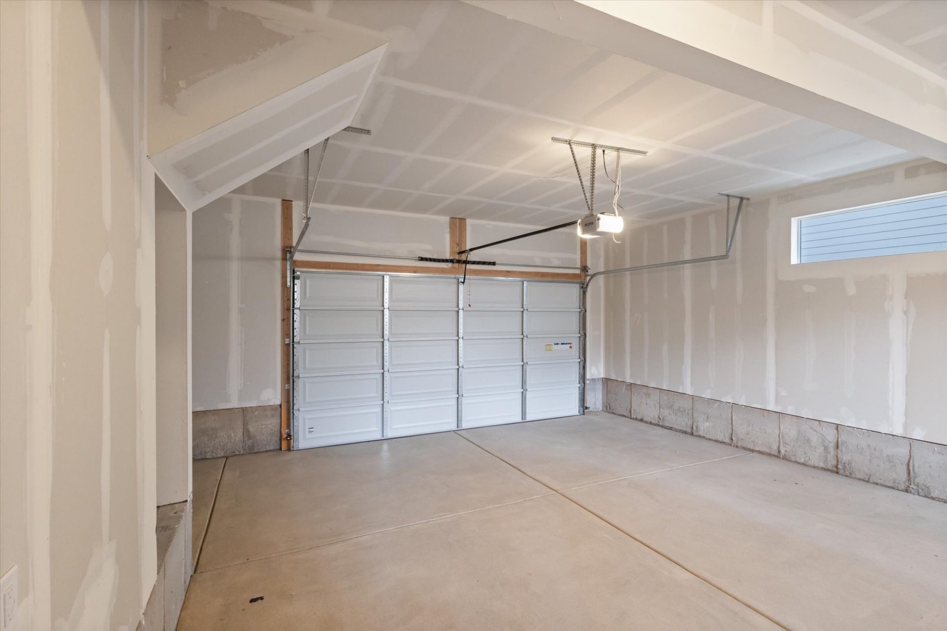 A clean, empty two-car garage with unfinished drywall walls, a concrete floor, bright overhead light, and a closed white sectional garage door with windows near the ceiling.
